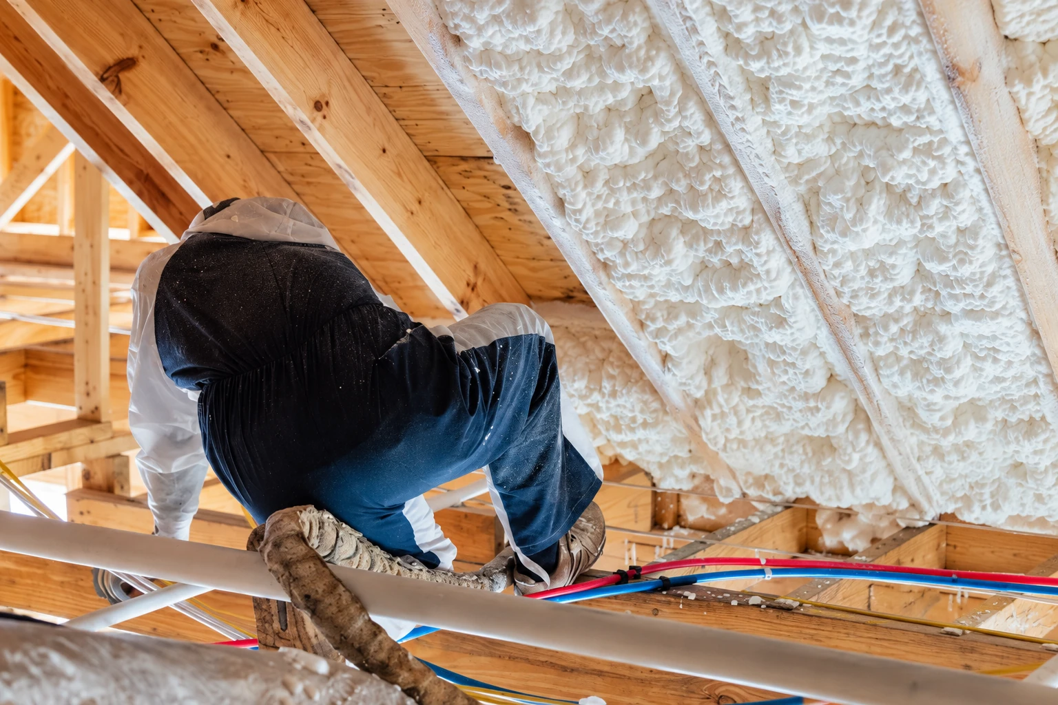 Worker applying spray foam insulation between exposed roof rafters in an unfinished attic.