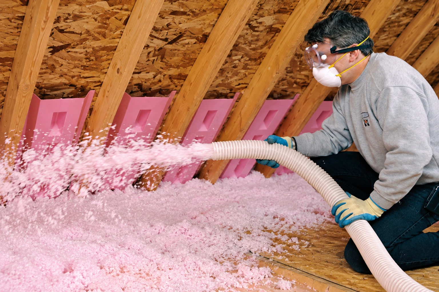 A worker wearing goggles and a respirator sprays pink blown-in insulation across an attic floor between roof rafters.