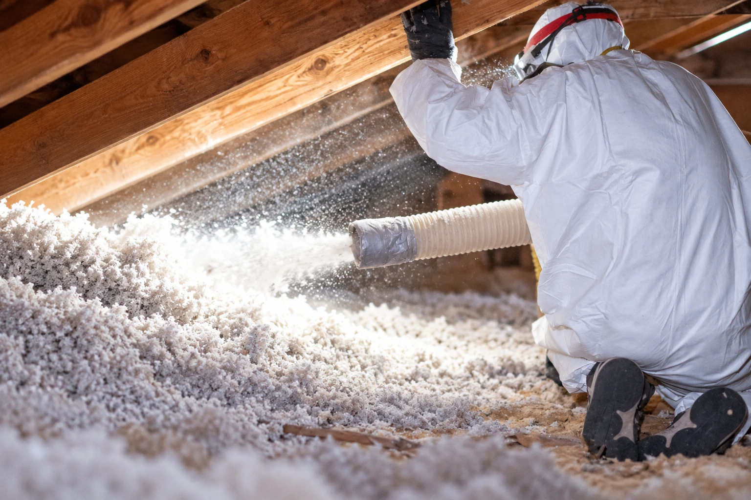 Worker in protective clothing sprays loose-fill insulation through a hose across an attic floor.