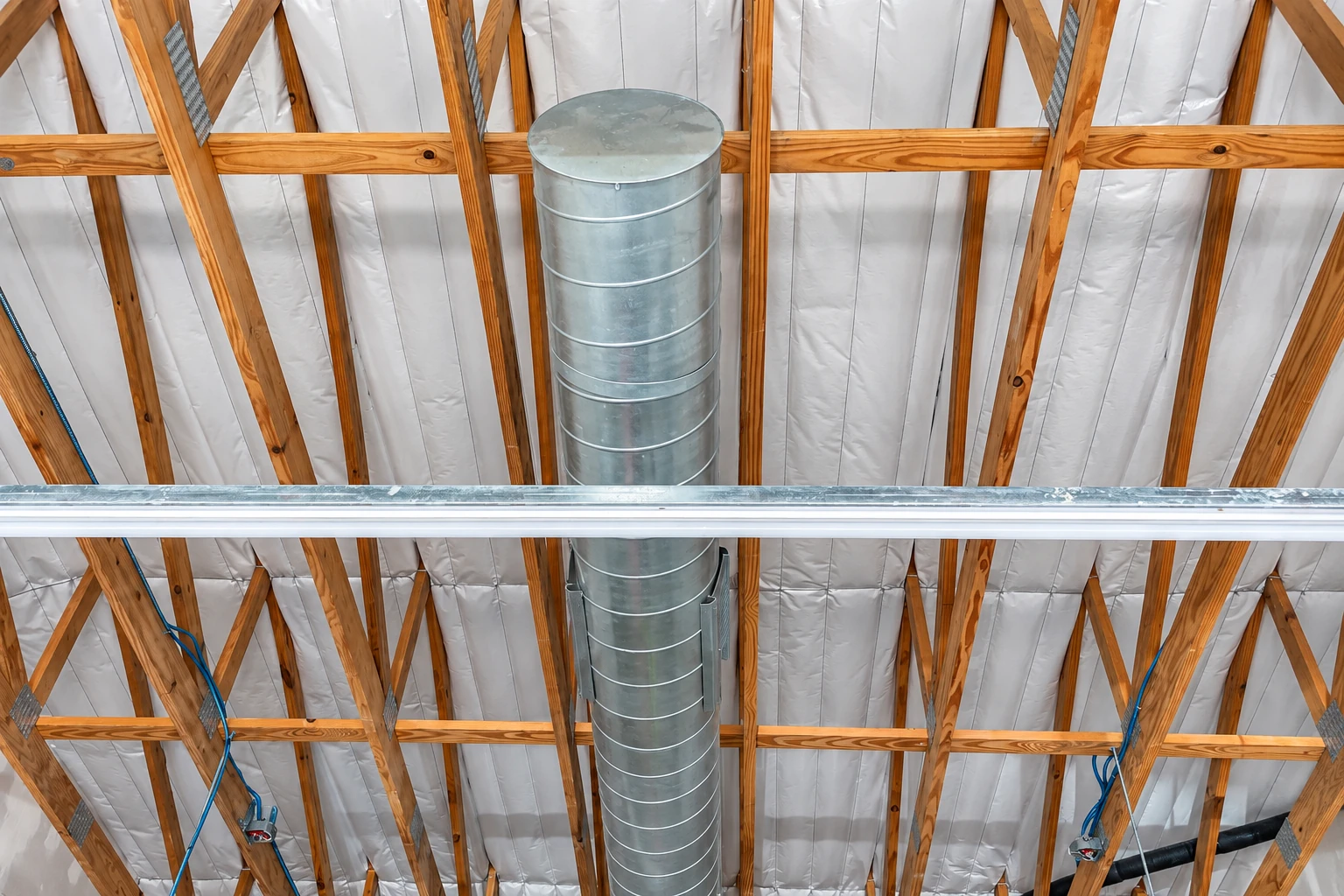 Commercial roof framing with white insulation panels between wooden trusses and a large central metal duct.