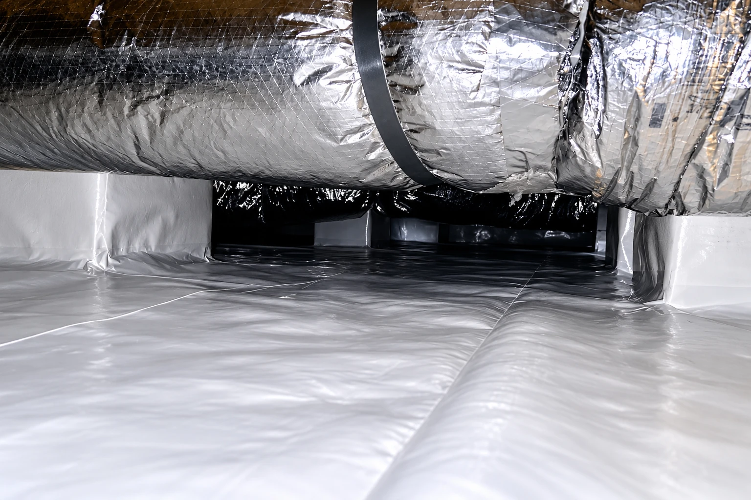 A crawl space has a smooth white vapor barrier across the floor and walls beneath a foil-wrapped duct.