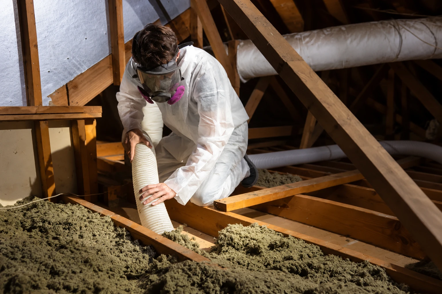 A worker in protective coveralls and a respirator removes loose attic insulation with a large vacuum hose.