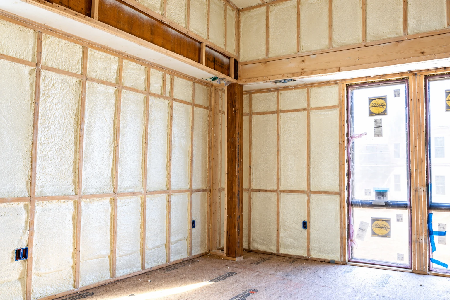 Spray foam insulation fills the wall cavities and upper framing of an unfinished room beside tall windows.